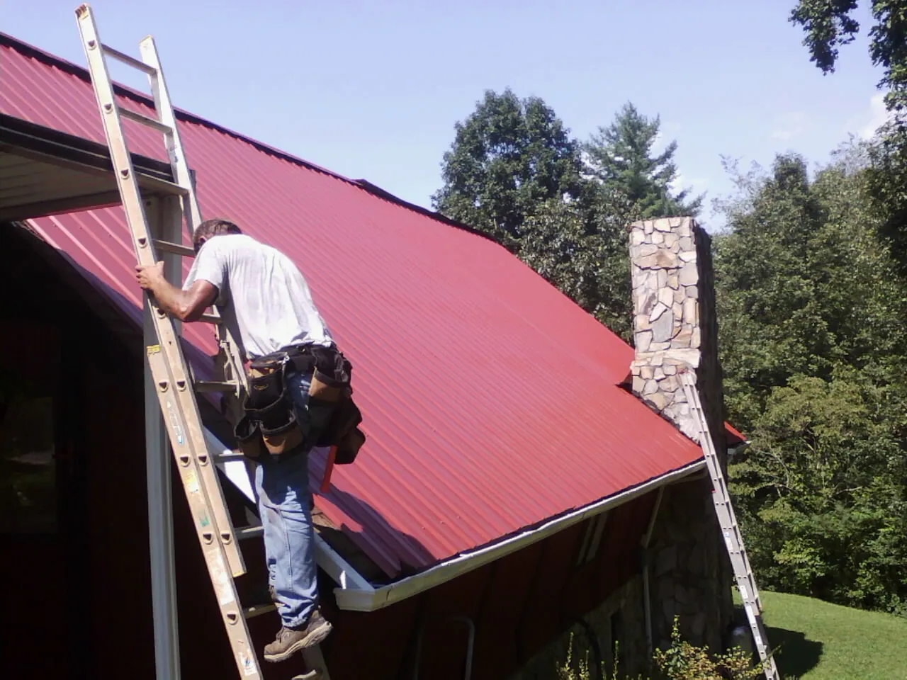 Roofer on a ladder beside a red metal roof and stone chimney — Soffit & Fascia Repair by Roofing Brothers in Terryville